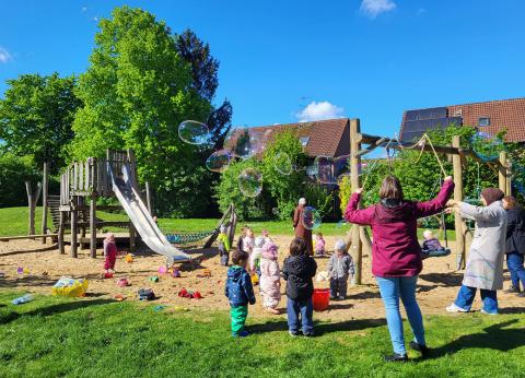 Mehrere Kinder und Erwachsene spielen auf einem Spielplatz mit Seifenblasen.